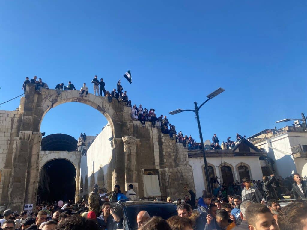 People celebrating in a town square, including standing on a rooftop with Syrian flags.